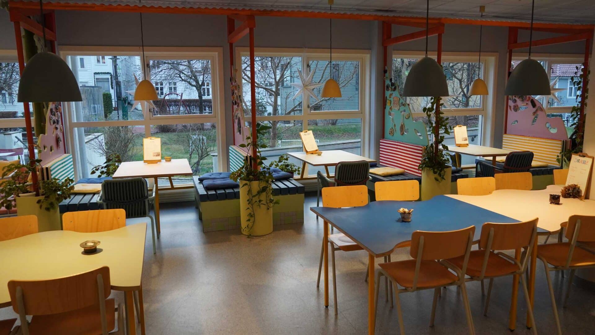 The colourful interior of Kafe Mat & prat. Several coloured tables and chair with hanging lights above them as well as some booths next to the windows overlooking the park outside.